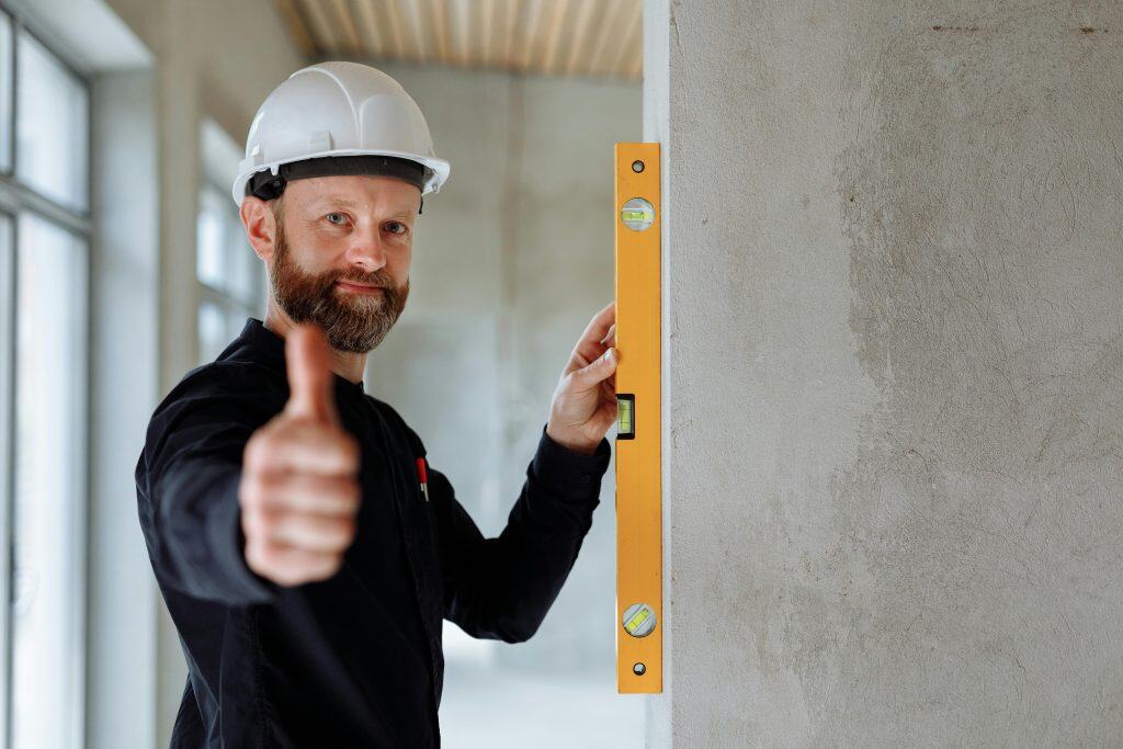 Professional construction worker with hard hat and spirit level giving thumbs up indoors.